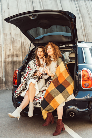 laughing adult women wrapped in blankets holding soda bottles sitting in car trunk at urban streetの写真素材