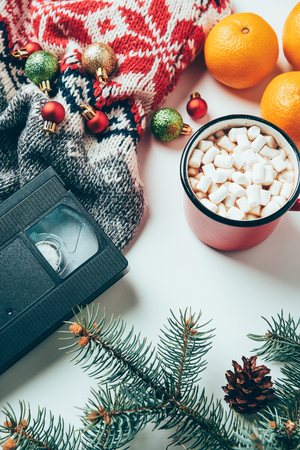Top view of video cassette, sweater, Christmas balls and cup of hot drink with marshmallows on white surfaceの写真素材