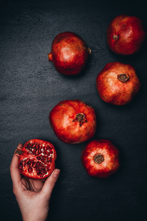 partial view of woman holding cut pomegranate on black surface with arranged wholesome garnetsの写真素材
