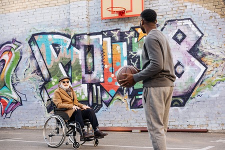 Happy senior disabled man in wheelchair and African american man playing basketball together on...