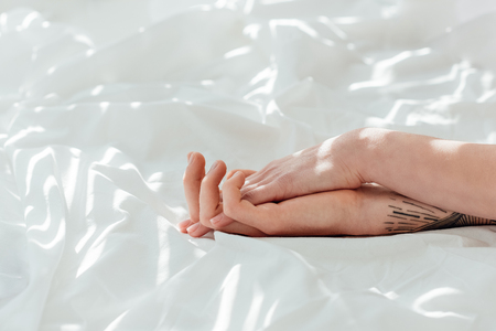 Partial view of loving couple holding hands while lying on white bed sheetの写真素材