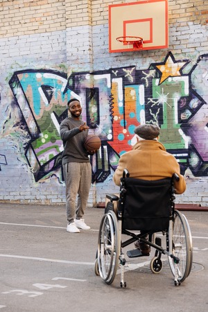Rear view of senior disabled man in wheelchair looking at happy African american man playing...