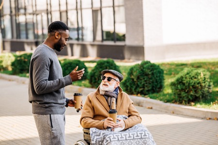 Smiling senior disabled man in wheelchair with plaid and African american man spending tome together on street with paper cups of coffeeの写真素材