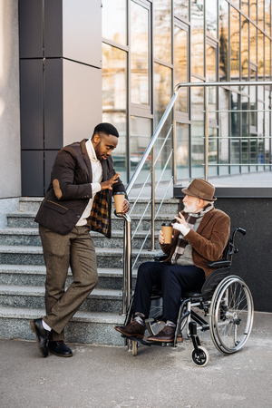 Senior disabled man in wheelchair and African american man drinking coffee together on streetの写真素材