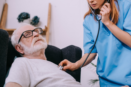 Nurse with stethoscope checking heartbeat of sick senior patientの写真素材