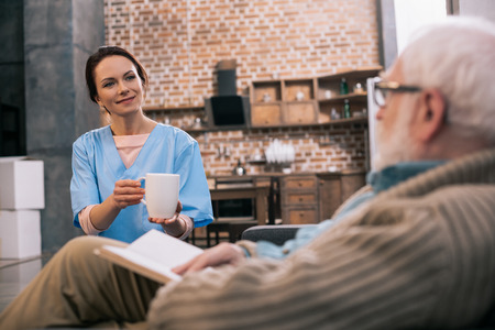 Nurse giving cup with drink to senior patient with bookの写真素材
