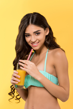 beautiful smiling girl posing in bikini with glass of juice, isolated on yellowの写真素材