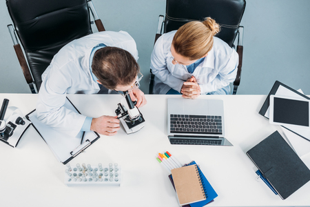 overhead view of scientific researchers in white coats working together at workplace with microscope and laptop in laboratoryの写真素材