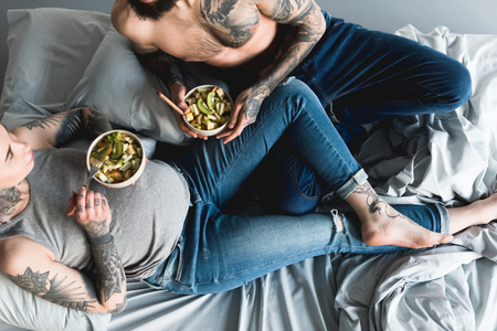 cropped image of pregnant tattooed girlfriend eating salad with boyfriend on bedの写真素材