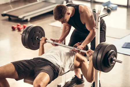 athletic trainer helping sportsman lifting barbell with heavy weight plates in gym