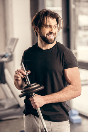 smiling handsome sportsman fixing weight plates on iron bar for training in gymの写真素材