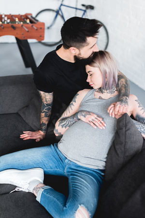 high angle view of tattooed boyfriend hugging pregnant girlfriend on sofa at homeの写真素材