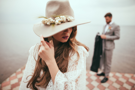 beautiful bride in wedding dress and hat in boho style on pier at lake, groom standing behindの写真素材