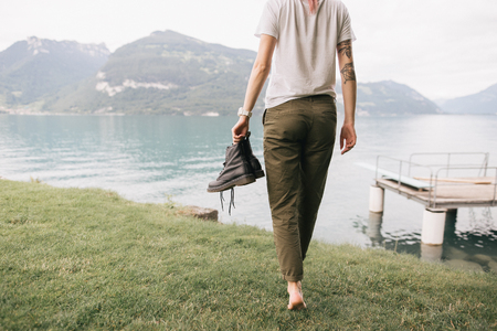 cropped shot of girl holding shoes and walking barefoot on grass near beautiful mountain lakeの写真素材