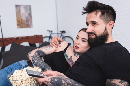 affectionate tattooed couple watching tv with popcorn at homeの写真素材
