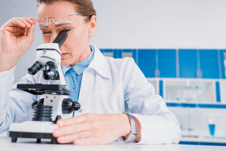 female scientist in lab coat and eyeglasses looking through microscope on reagent in labの写真素材