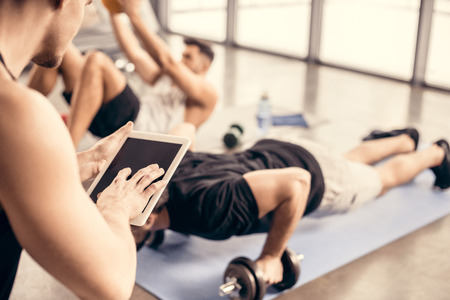 trainer using tablet and counting results of sportsman doing push ups on dumbbells in gym