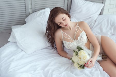 sexy woman in white underwear with bouquet of roses resting on bedの写真素材