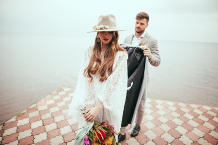 attractive bride in wedding dress and hat and groom on pier at lakeの写真素材