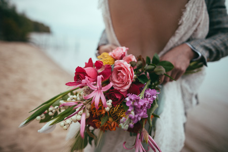 cropped view of bride and groom holding wedding bouquet and embracing on beachの写真素材