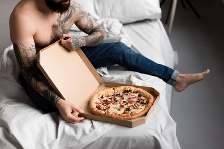 cropped image of shirtless tattooed man with pizza on bedの写真素材