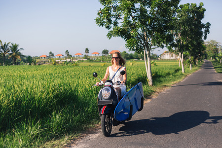 young smiling woman riding scooter with surfing boardの写真素材