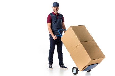 smiling delivery man in uniform pushing hand truck with cardboard boxes isolated on whiteの写真素材