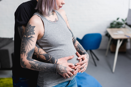 cropped image of tattooed boyfriend hugging pregnant girlfriend from back in living roomの写真素材