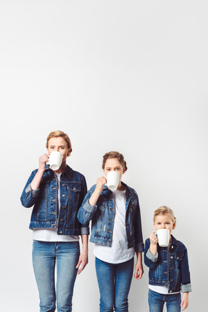 portrait of mother and daughters in similar denim clothing drinking hot drinks isolated on greyの写真素材