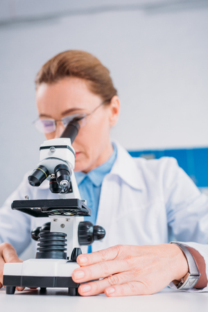 selective focus of female scientist in lab coat and eyeglasses looking through microscope on reagent in labの写真素材