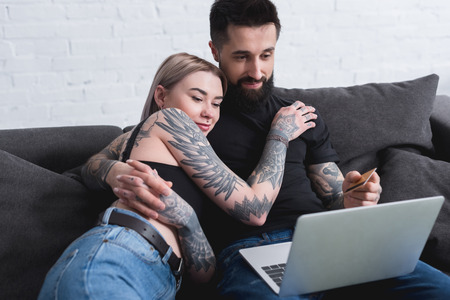 happy tattooed couple looking at laptop on sofa at homeの写真素材