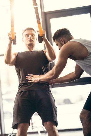 trainer helping young sportsman exercising with suspension straps in gymの写真素材