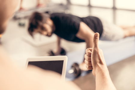 trainer holding tablet and showing thumb up to sportsman doing push ups on dumbbells in gymの写真素材