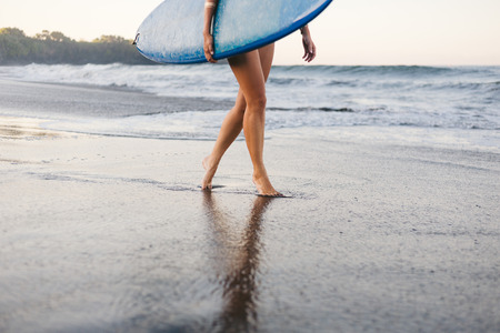 cropped shot of sportswoman in swimming suit with blue surfing board walking on coastlineの写真素材