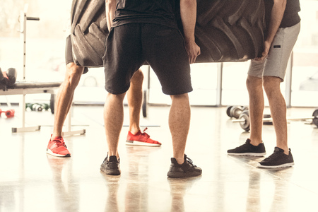 low section of three athletic men in sportswear lifting tyre in gym