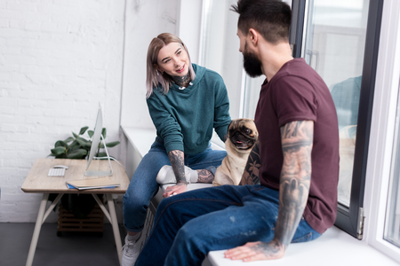 happy tattooed couple sitting on windowsill with pug at homeの写真素材
