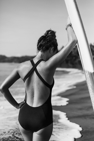 back view of young girl in swimsuit posing with surfboard near ocean, black and whiteの写真素材
