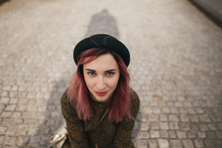 young girl with pink hair in trendy hat sitting on pavement in cityの写真素材