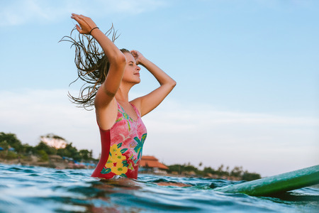 young woman readjusting wet hair while sitting on surfing board in oceanの写真素材