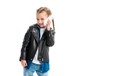 Smiling stylish kid wearing leather jacket and talking on smartphone isolated on whiteの写真素材