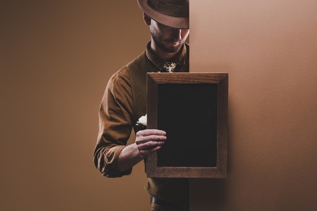 Stylish man in hat holding empty frame isolated on brownの写真素材
