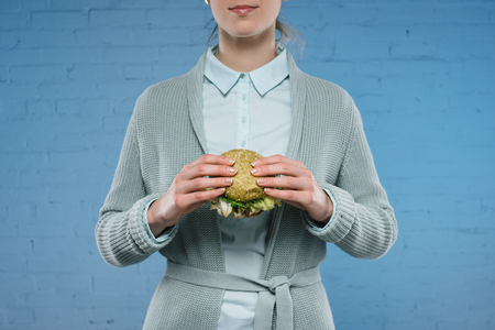cropped shot of young woman holding green vegan burger in front of blue brick wallの写真素材