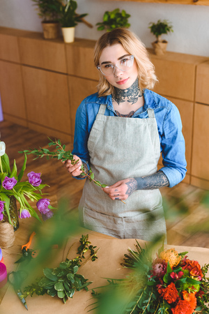 beautiful young florist in apron and eyeglasses arranging flowers and looking at cameraの写真素材