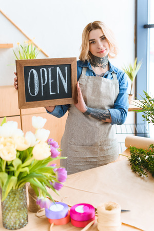 beautiful young florist holding open sign and looking at camera in flower shopの写真素材