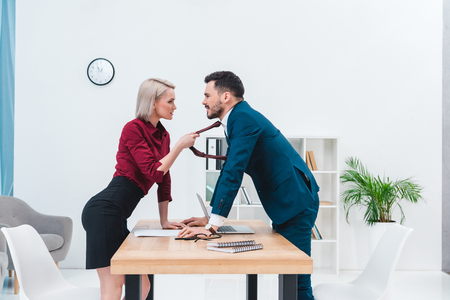 side view of young businesswoman holding necktie of handsome business man and flirting in officeの写真素材