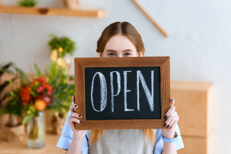 young female florist holding open sign and looking at camera in flower shopの写真素材