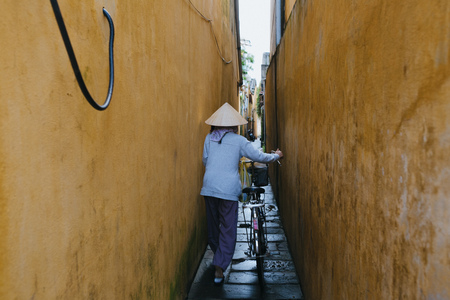 HOI AN, VIETNAM - 03 JANUARY, 2018: back view of vietnamese woman in traditional hat carrying bicycle on narrow street in Hoi An, Vietnamのeditorial素材