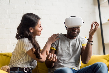 smiling african american woman sitting near expressive boyfriend while he wearing virtual reality...