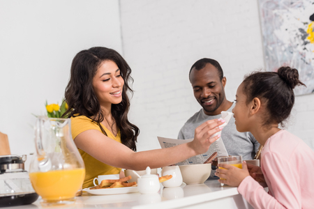 happy mother wiping face of little daughter after breakfast while father reading newspaper on kitchenの写真素材