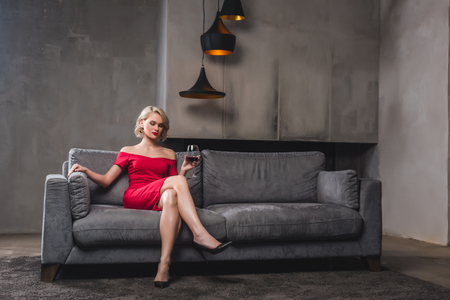 beautiful blonde woman in red dress sitting on sofa and drinking wineの写真素材